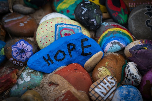 A collection of painted stones with various designs. A prominent blue stone features the word 'HOPE' in black letters. Other stones showcase colorful patterns, including a rainbow, a flower with '2020' written alongside, and an NHS logo. The stones are arranged closely together on a sandy surface.