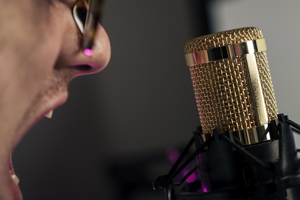 Close-up of a person, likely singing or speaking, with an open mouth near a gold-colored microphone setup on a stand. The person's face is partially visible, focusing on the mouth and nose, with glasses visible on the person's face.