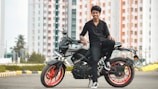 A young person in black clothing is sitting on a modern motorcycle with red rims. The background features high-rise buildings with a pattern of red and white, along with trees and an urban landscape. The scene conveys a sense of style and modernity.