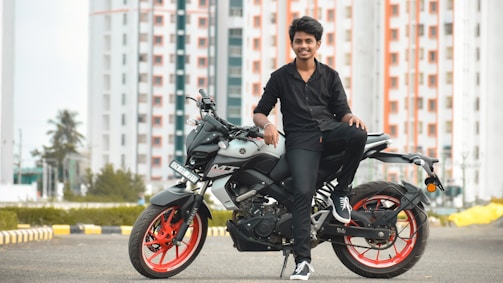 A young person in black clothing is sitting on a modern motorcycle with red rims. The background features high-rise buildings with a pattern of red and white, along with trees and an urban landscape. The scene conveys a sense of style and modernity.