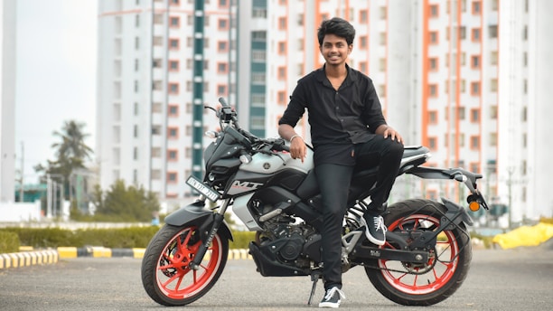 A young person in black clothing is sitting on a modern motorcycle with red rims. The background features high-rise buildings with a pattern of red and white, along with trees and an urban landscape. The scene conveys a sense of style and modernity.