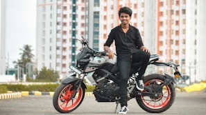 A young person in black clothing is sitting on a modern motorcycle with red rims. The background features high-rise buildings with a pattern of red and white, along with trees and an urban landscape. The scene conveys a sense of style and modernity.