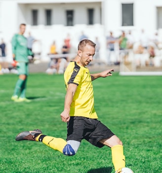 boy in yellow and black soccer jersey kicking soccer ball on green grass field during daytime