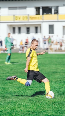 boy in yellow and black soccer jersey kicking soccer ball on green grass field during daytime