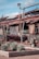 An industrial scene with an old, rusted structure and broken-down machinery. The roof is partially collapsed and scattered debris surrounds the area. Yucca plants are arranged in a landscaped area with reddish mulch. Overhead lights are mounted on poles, and the sky is clear with a few clouds.