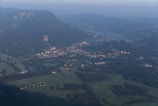 A dense, verdant landscape with a sprawling town nestled at the base of a large, forested mountain. The town is surrounded by lush fields and a winding river that snakes through the landscape. Several buildings, roads, and patches of farmland are visible, indicating a well-established community.