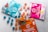 A variety of cannabis-infused gummy packages are displayed on a light surface. Three distinct packages in blue, pink, and orange colors are each accompanied by spherical gummies in matching hues. The blue package features illustrations of berries, while the pink one shows a lemon graphic. The orange package is labeled as vegan.