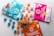 A variety of cannabis-infused gummy packages are displayed on a light surface. Three distinct packages in blue, pink, and orange colors are each accompanied by spherical gummies in matching hues. The blue package features illustrations of berries, while the pink one shows a lemon graphic. The orange package is labeled as vegan.