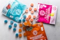 A variety of cannabis-infused gummy packages are displayed on a light surface. Three distinct packages in blue, pink, and orange colors are each accompanied by spherical gummies in matching hues. The blue package features illustrations of berries, while the pink one shows a lemon graphic. The orange package is labeled as vegan.