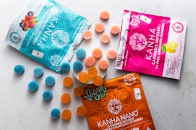 A variety of cannabis-infused gummy packages are displayed on a light surface. Three distinct packages in blue, pink, and orange colors are each accompanied by spherical gummies in matching hues. The blue package features illustrations of berries, while the pink one shows a lemon graphic. The orange package is labeled as vegan.