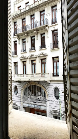 A view through an open window toward a classic European-style building with ornate architecture. The building features detailed columns, decorative railings on the balconies, and intricate stonework. The window shutters are partially visible, framing the scene.