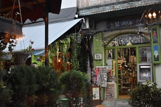 A quaint shopfront adorned with various hanging plants and decorative lights. The store has a rustic feel, with green-framed windows displaying an assortment of colorful trinkets and souvenirs. There are plants outside, including bushes, and a hanging basket. On the right, a small ATM is embedded into the wall.