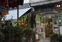 A quaint shopfront adorned with various hanging plants and decorative lights. The store has a rustic feel, with green-framed windows displaying an assortment of colorful trinkets and souvenirs. There are plants outside, including bushes, and a hanging basket. On the right, a small ATM is embedded into the wall.
