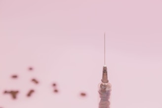 A single syringe needle stands upright against a soft pink background with several small, blurry objects scattered in the distance.