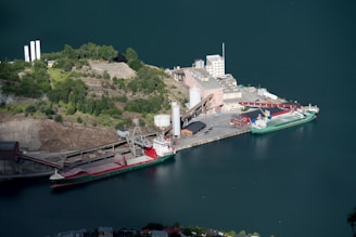 Aerial view of a port facility featuring a docked cargo ship. The surrounding area includes industrial buildings and a lush, green landscape. The deep blue water contrasts with the green hills. Industrial structures like silos and warehouses are visible, along with a narrow, winding road leading to the port.