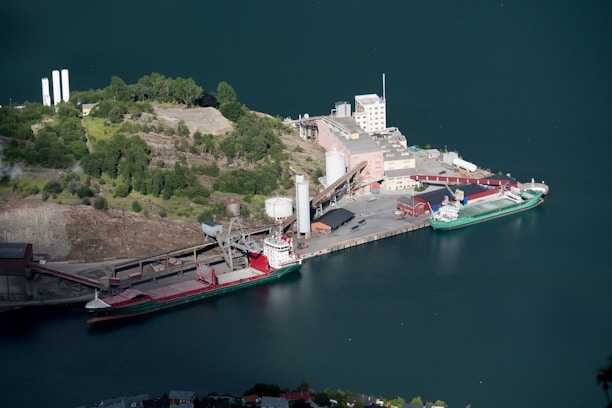 Aerial view of a port facility featuring a docked cargo ship. The surrounding area includes industrial buildings and a lush, green landscape. The deep blue water contrasts with the green hills. Industrial structures like silos and warehouses are visible, along with a narrow, winding road leading to the port.