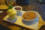 A wooden table hosts a dessert spread featuring a small glass of vanilla ice cream garnished with fresh mint leaves, a bowl of rich chocolate sauce, and a baked chocolate dessert in a white ceramic dish. The items are arranged on a wooden serving board lined with a piece of branded parchment paper.