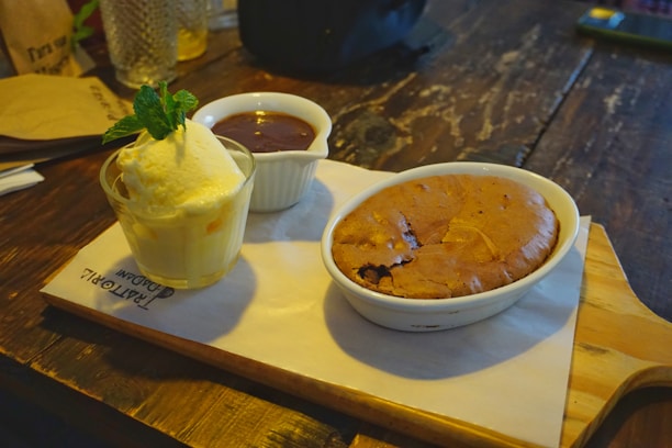 A wooden table hosts a dessert spread featuring a small glass of vanilla ice cream garnished with fresh mint leaves, a bowl of rich chocolate sauce, and a baked chocolate dessert in a white ceramic dish. The items are arranged on a wooden serving board lined with a piece of branded parchment paper.