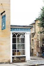 A cozy street corner features a small building with a sign that reads 'Cafe Loco'. The facade includes a large window with an arched design, revealing a wicker chair inside. The building is surrounded by cobblestone paths and nearby structures made of brick and stone. A bicycle is seen parked on the right, and a person stands nearby.