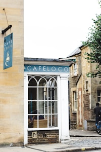 A cozy street corner features a small building with a sign that reads 'Cafe Loco'. The facade includes a large window with an arched design, revealing a wicker chair inside. The building is surrounded by cobblestone paths and nearby structures made of brick and stone. A bicycle is seen parked on the right, and a person stands nearby.