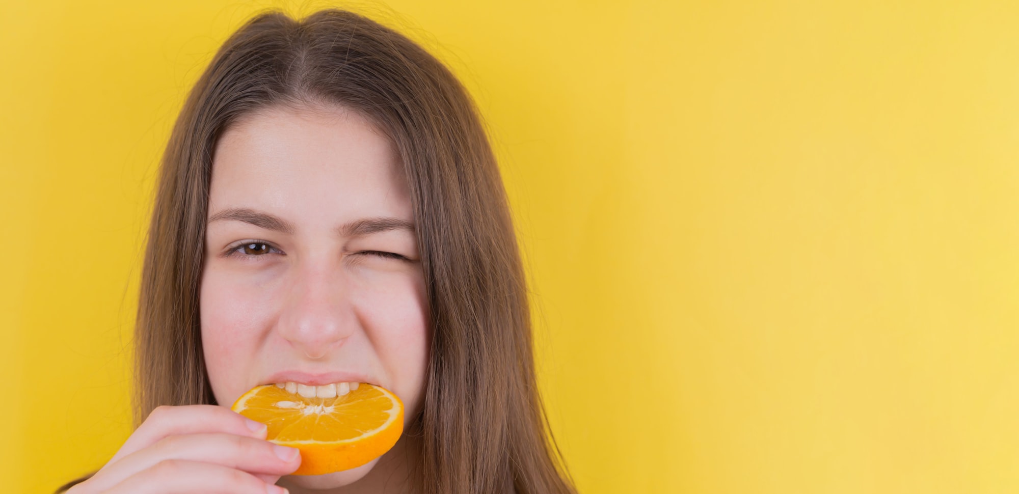 A cute brunette girl on a yellow background bites a slice of sour orange and winks. Place for an inscription. Fruits. Sour. Close plan. Advertising.