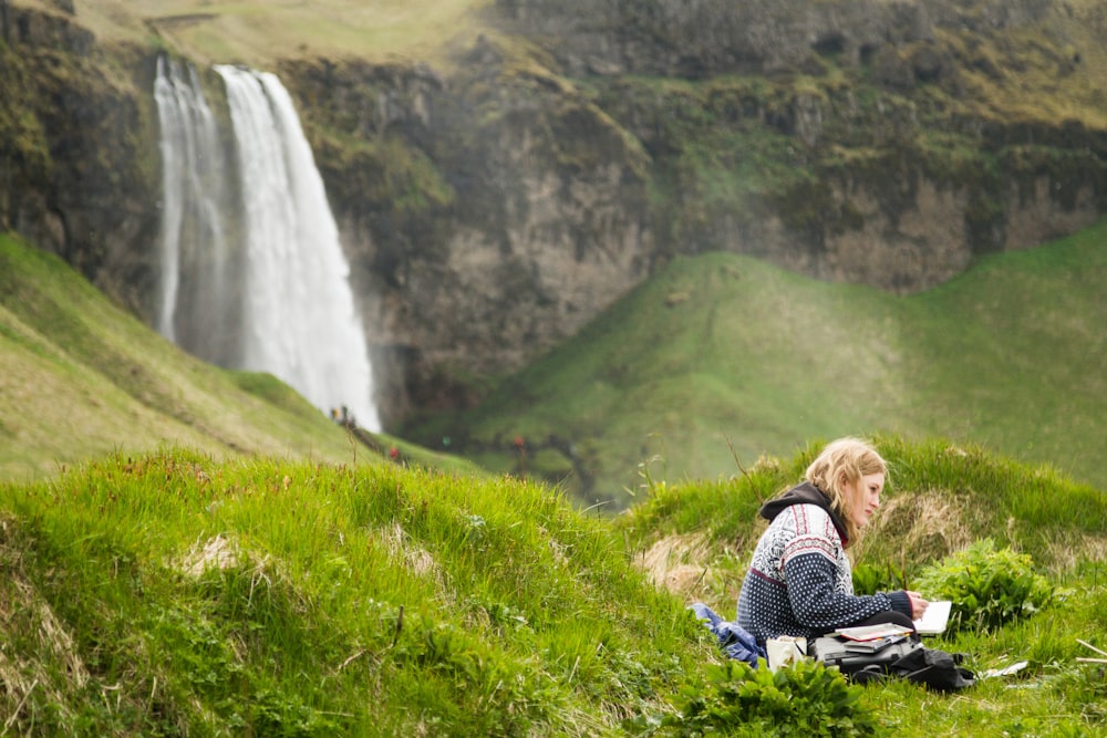 woman in black and white jacket sitting on green grass field near waterfalls during daytime
