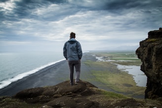 man in gray jacket standing on brown rock near body of water during daytime