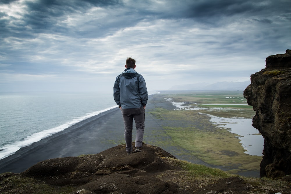 man in gray jacket standing on brown rock near body of water during daytime