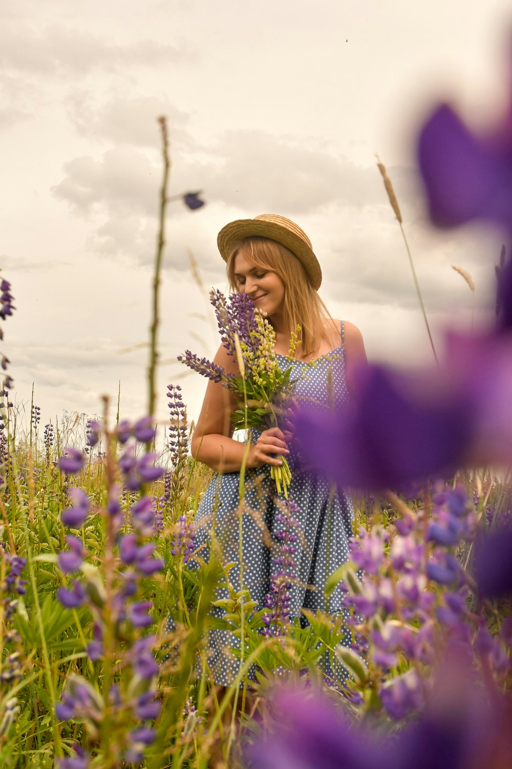 woman in purple and white polka dot dress wearing brown hat standing on purple flower field