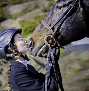 A person wearing a riding helmet and braided hair is affectionately kissing the nose of a horse. The person is dressed in equestrian gear and appears to be holding the bridle of the horse. The background consists of a blurred natural landscape with green foliage.