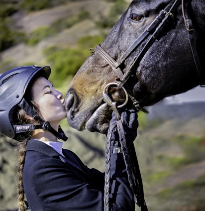 A person wearing a riding helmet and braided hair is affectionately kissing the nose of a horse. The person is dressed in equestrian gear and appears to be holding the bridle of the horse. The background consists of a blurred natural landscape with green foliage.