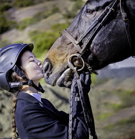 A person wearing a riding helmet and braided hair is affectionately kissing the nose of a horse. The person is dressed in equestrian gear and appears to be holding the bridle of the horse. The background consists of a blurred natural landscape with green foliage.