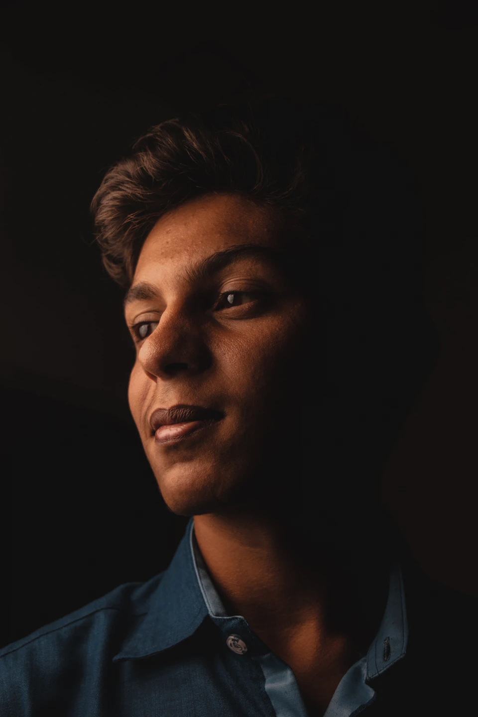 The image shows a young man with brown hair wearing a blue collared shirt, standing against a dark background and looking to his left side. The man's gaze is directed off to the side, suggesting he may be lost in thought or observing something outside of the frame.