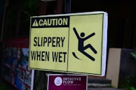 A yellow caution sign with black text that reads 'Caution Slippery When Wet' and an illustration of a person slipping. Below it, there is a sign for 'Intuitive Flow Yoga Studio' with a symbol and text indicating its location in Ubud, Bali.