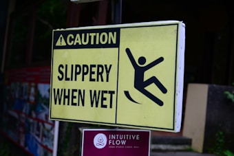 A yellow caution sign with black text that reads 'Caution Slippery When Wet' and an illustration of a person slipping. Below it, there is a sign for 'Intuitive Flow Yoga Studio' with a symbol and text indicating its location in Ubud, Bali.