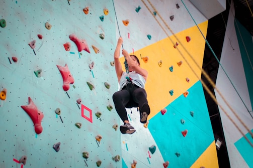 A person is climbing an indoor rock wall, which is equipped with colorful holds of various shapes and sizes. The climber is wearing a harness and appears to be mid-climb, showcasing determination and focus.