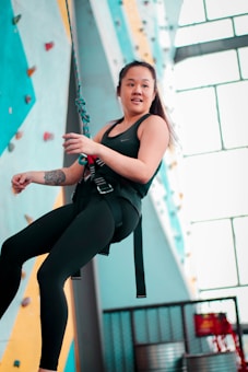 A person is engaged in indoor rock climbing, secured with a rope and wearing climbing gear. The colorful climbing wall features various holds and the setting is illuminated by natural light coming through large windows.