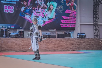 A person in elaborate costume, possibly for a cosplay event, stands in front of a stage. The stage has musical equipment like speakers and amplifiers. A large banner in the background displays the text 'Click Youth Festival 2018' with colorful graphics and is surrounded by images of characters. The floor is multi-colored, suggesting an indoor or exhibition space.