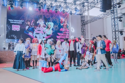 A group of people dressed in various colorful costumes, likely participating in a cosplay event. The backdrop features a large banner with graphics of characters and information about a youth festival. The setting appears to be indoors with a stage and lighting fixtures above.