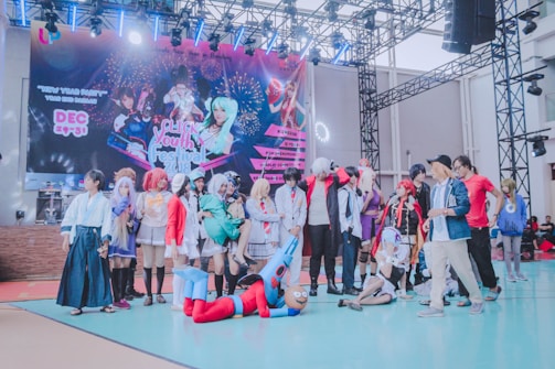A group of people dressed in various colorful costumes, likely participating in a cosplay event. The backdrop features a large banner with graphics of characters and information about a youth festival. The setting appears to be indoors with a stage and lighting fixtures above.
