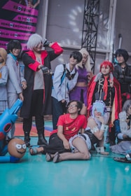 A group of people are dressed in various cosplay costumes, posing together in a festive environment. The costumes range from bright red to dark hues, with various accessories like wigs and props enhancing their outfits. Some individuals are standing, while others are kneeling on the ground, creating a lively and engaging scene. The background includes a colorful banner with text.