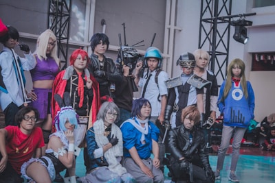 A group of people in elaborate and colorful costumes standing together, likely participating in a cosplay event. Each individual appears to be dressed as a distinct character, featuring various accessories such as wigs, masks, armor, and themed clothing. The setting is indoors, with lighting and staging equipment visible.