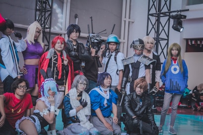 A group of people in elaborate and colorful costumes standing together, likely participating in a cosplay event. Each individual appears to be dressed as a distinct character, featuring various accessories such as wigs, masks, armor, and themed clothing. The setting is indoors, with lighting and staging equipment visible.