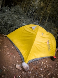 A yellow tent is pitched on a dirt surface surrounded by dense, green forest. The tent is marked with a brand name and has rocks at its base to secure it. Debris like small rocks and litter are scattered on the ground.