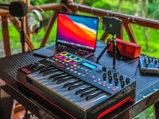 A music production setup placed on a wooden porch features a MIDI keyboard with control knobs, a laptop displaying vivid colors on its screen, a microphone on a stand, headphones hanging on a railing, a portable speaker, and a mixer. The background shows lush greenery, adding a serene ambiance.