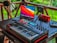A music production setup placed on a wooden porch features a MIDI keyboard with control knobs, a laptop displaying vivid colors on its screen, a microphone on a stand, headphones hanging on a railing, a portable speaker, and a mixer. The background shows lush greenery, adding a serene ambiance.