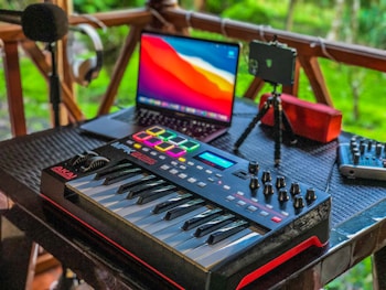 A music production setup placed on a wooden porch features a MIDI keyboard with control knobs, a laptop displaying vivid colors on its screen, a microphone on a stand, headphones hanging on a railing, a portable speaker, and a mixer. The background shows lush greenery, adding a serene ambiance.