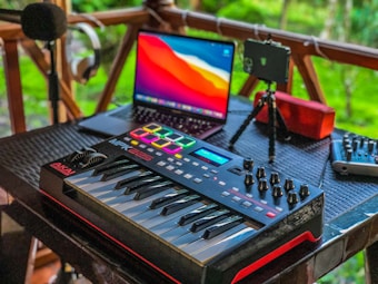 A music production setup placed on a wooden porch features a MIDI keyboard with control knobs, a laptop displaying vivid colors on its screen, a microphone on a stand, headphones hanging on a railing, a portable speaker, and a mixer. The background shows lush greenery, adding a serene ambiance.