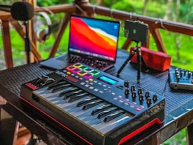A music production setup placed on a wooden porch features a MIDI keyboard with control knobs, a laptop displaying vivid colors on its screen, a microphone on a stand, headphones hanging on a railing, a portable speaker, and a mixer. The background shows lush greenery, adding a serene ambiance.