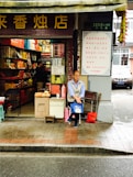 A street-side shop with an open front showcases a variety of colorful packaged goods inside. A man is seated at the entrance, holding a blue bag, with additional items like a red bag and cardboard boxes nearby. The storefront has signage with Chinese characters, and the surrounding environment appears to be urban with a road in the foreground.